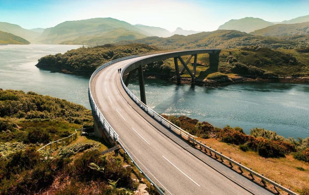 A curving bridge spans over a serene river, surrounded by lush green hills and distant mountains under a clear blue sky.