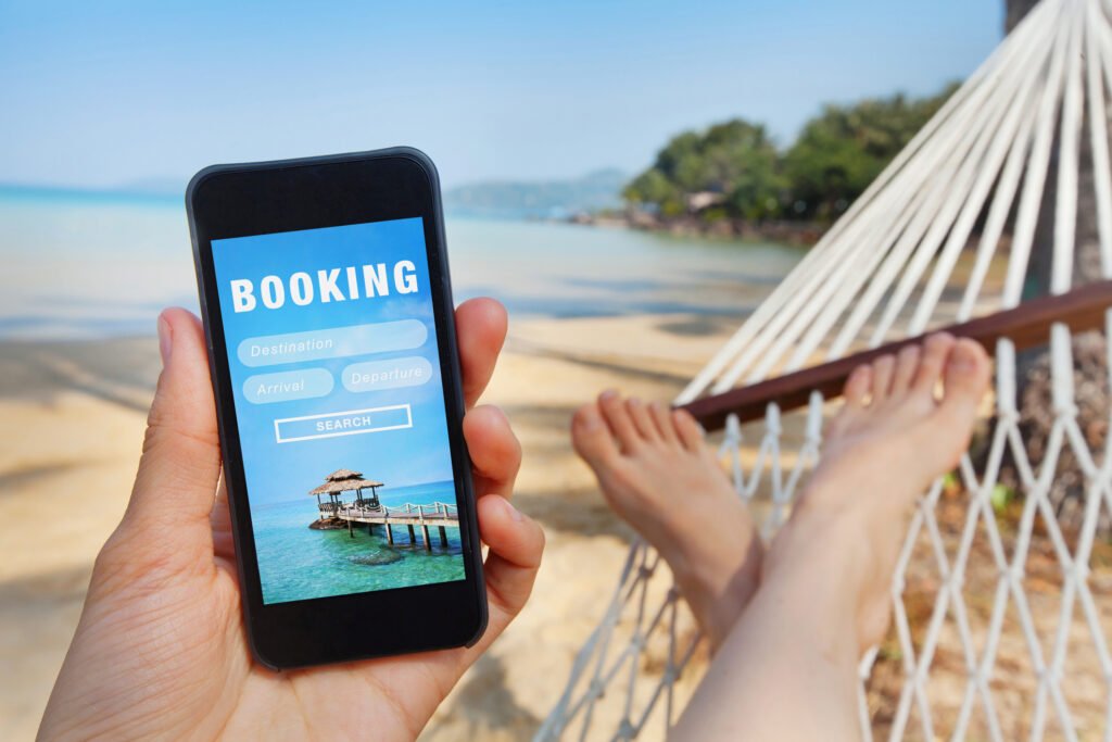 A person looks at a phone with a travel "booking" screen while lying on a hammock on a beach.