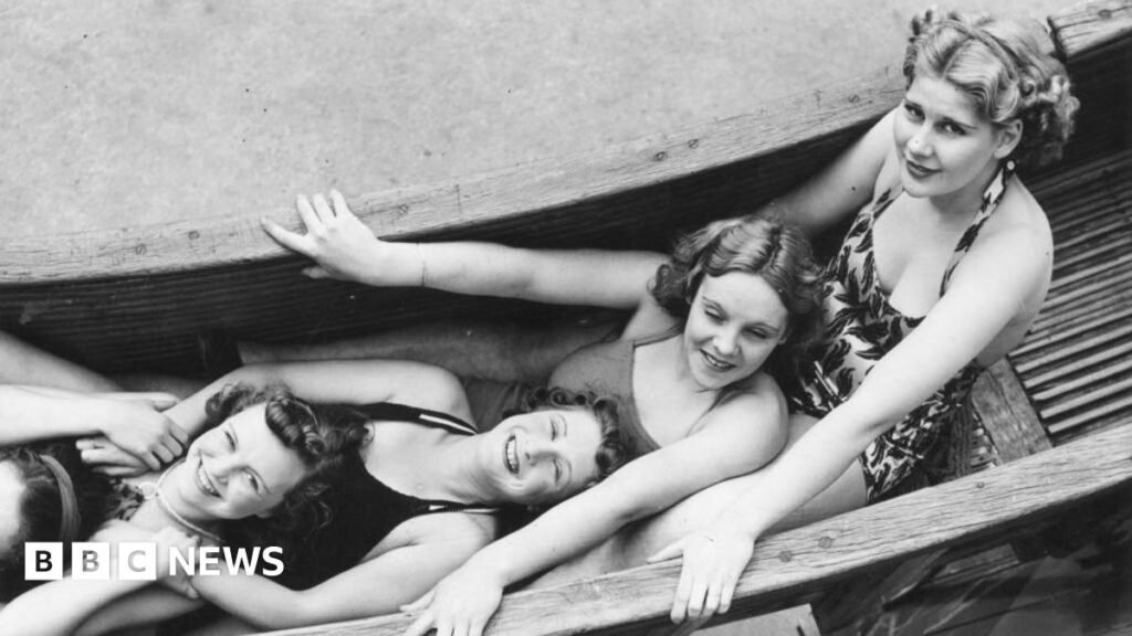 A black and white photo of four women wearing swimwear, lying down in a row on a wooden helterskelter ride and smiling at the camera.