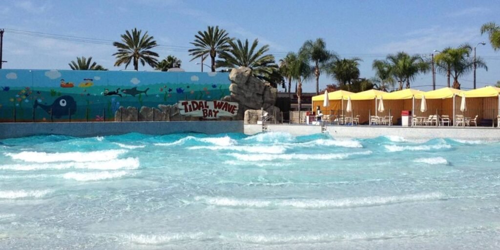 A wave pool with turquoise water and small waves, labeled "Tidal Wave Bay" at Soak City. Palm trees, a mural with sea animals, yellow umbrellas, and white chairs decorate the background under a blue sky.