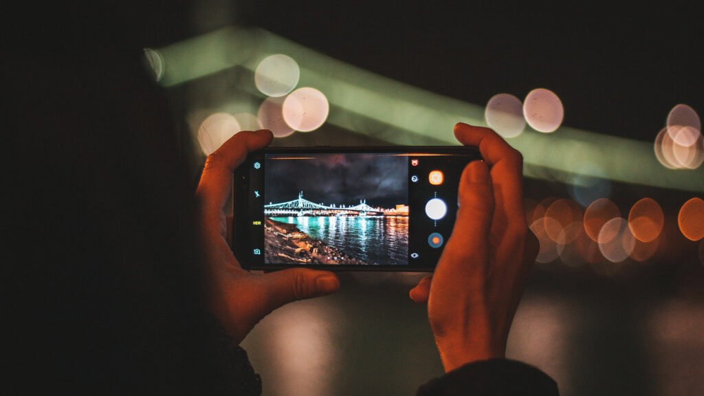 A person holds a smartphone, capturing a brightly lit bridge at night with colorful bokeh lights reflecting on the water