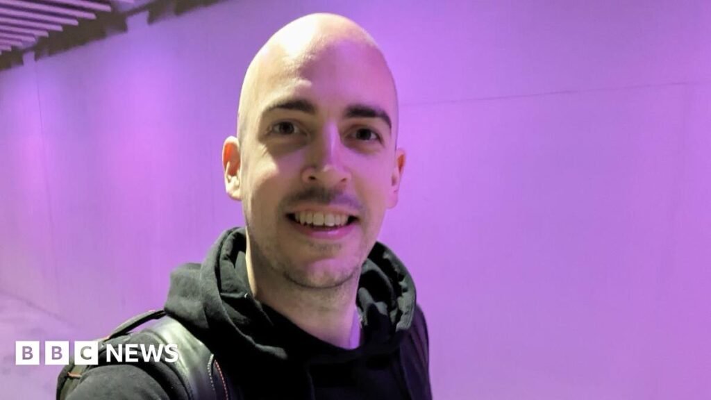 Dean Stokes - a man with no hair smiles into the camera. He has a black hoodie on and has a black bag over his shoulder.