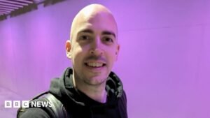 Dean Stokes - a man with no hair smiles into the camera. He has a black hoodie on and has a black bag over his shoulder.