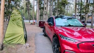 A red Ford Mustang Mach-E is parked in a forest campsite near a green tent. A person sits by a fire pit in the background, surrounded by trees and cabins. The scene feels peaceful and rustic.