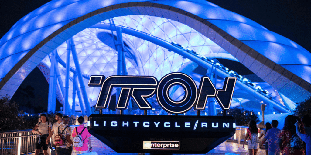 People walk toward the illuminated entrance of the TRON Lightcycle / Run attraction at night, beneath a glowing blue canopy and prominent TRON sign—an iconic scene often featured in any TRON versus Hagrid comparison guide at Magic Kingdom.
