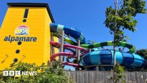 A yellow building with a logo reading Alpamare on the side. Several brightly coloured tube waterslides are exiting the side of the building and weaving around each other.