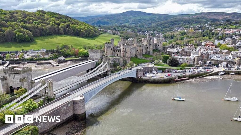 An aerial view shows Conwy Castle, on the banks of the River Conwy in north Wales - it is a bright day with clouds in the sky and the castle is at the forefront of the photo, connected across the river by a road and a footbridge. Conwy can be seen in the background and there are sail boats on the river heading towards the bridge from right to left. Behind and to the left of the town are rolling hills studded with trees.