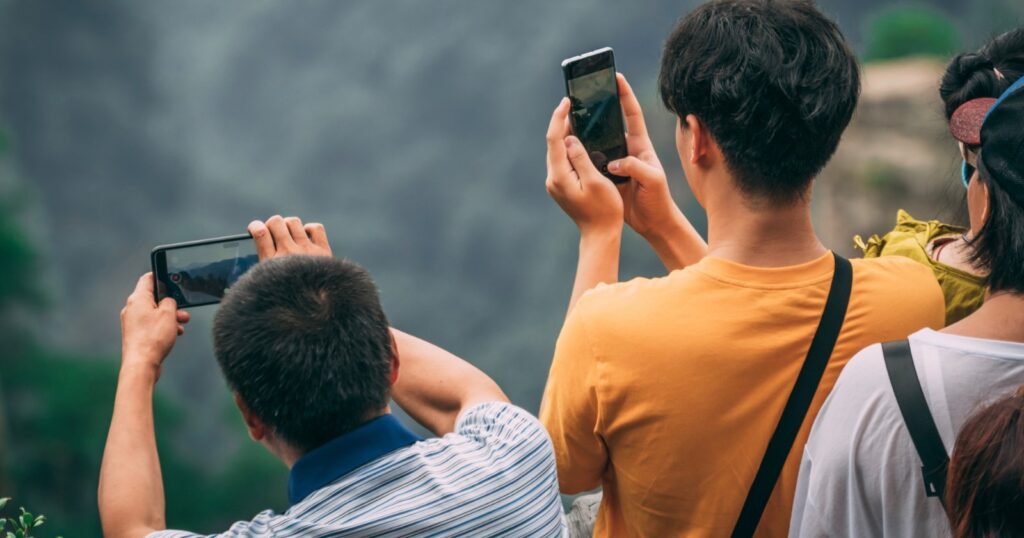 Three people standing outdoors, facing away from the camera, holding up smartphones to take photos of a natural landscape with blurred greenery and mountains in the background.
