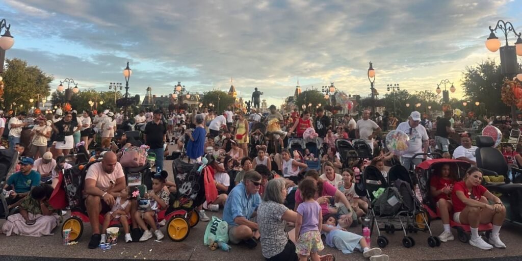 A large crowd of people, including families with children and strollers, sit and stand closely together outdoors under a cloudy sky, appearing to wait for an event at a theme park. Lamp posts and trees are in the background.