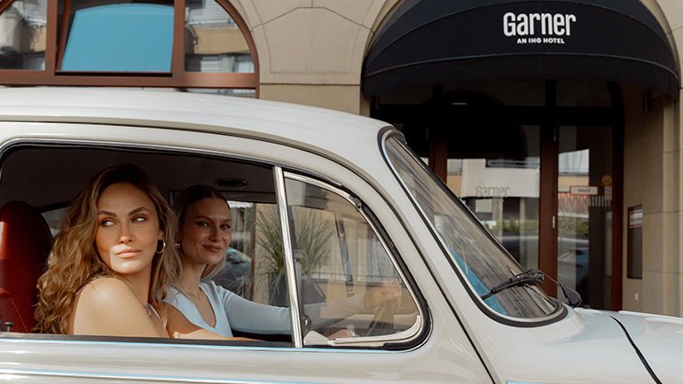 A side view of a light-colored vintage car with two women inside, parked in front of a hotel. The hotel entrance has a black awning with "Garner AN IHG HOTEL" written in white. The woman in the passenger seat, with long wavy hair, is looking to the side with a smile. The woman in the driver's seat is also smiling and looking forward.