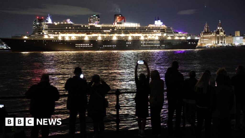 Night-time view of the ocean liner Queen Mary 2 on the river with the Liver building and Pier Head behind. About a dozen people can be seen in the foreground on the other side of the river taking pictures and watching.
