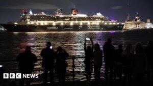 Night-time view of the ocean liner Queen Mary 2 on the river with the Liver building and Pier Head behind. About a dozen people can be seen in the foreground on the other side of the river taking pictures and watching.