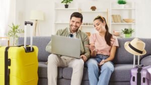 Newlywed couple planning their honeymoon trip, comparing prices on website while choosing tour online, enjoying summer holidays, posing on sofa with packed suitcase around. Travel concept
