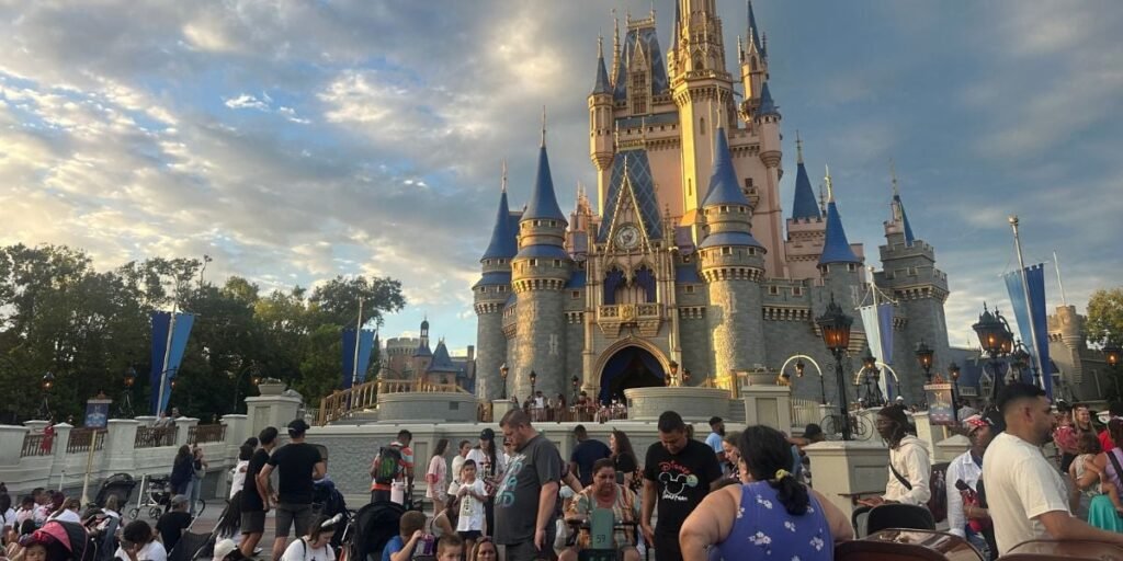 A large crowd of people gathered in front of Cinderella’s Castle at a Disney theme park, with the castle lit by the setting sun and a partly cloudy sky overhead.