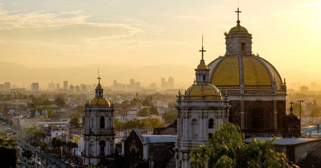 A scenic view of Mexico City at sunset, featuring the domes of a historic church in the foreground with modern city buildings and a hazy sky in the background.