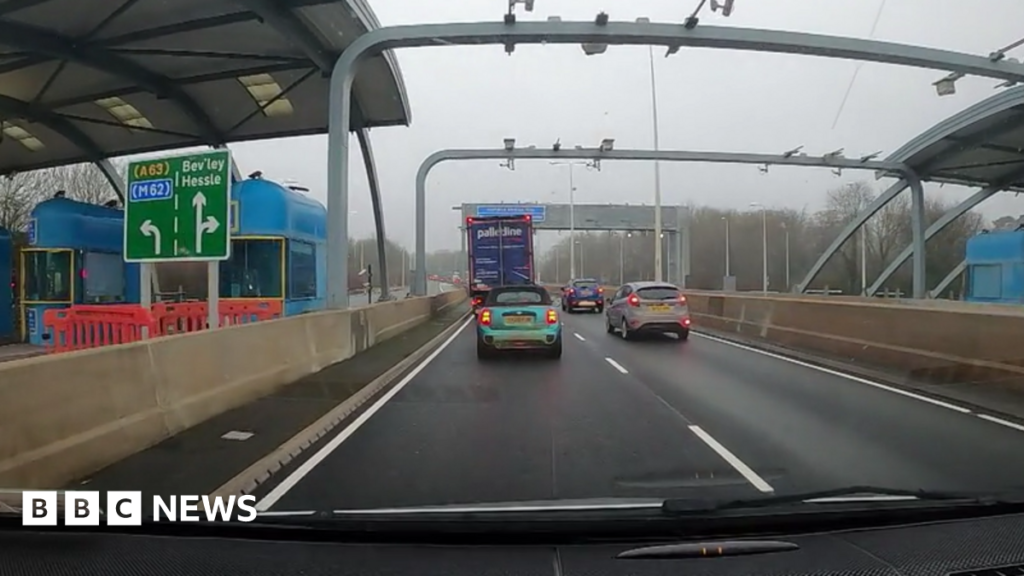 A man with short fair hair looks into the camera as he stands in front of toll booths at the entrance to a bridge. He is wearing a hi-vis orange jacked over a suit, lilac shirt and dark tie. Behind him is a red and white barrier blocking a road. A blue toll booth and red lights can also be seen. A truck is waiting at the toll booth. The sun is low in the sky.