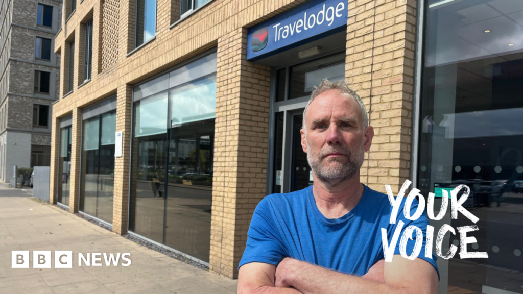 A man in a blue T-shirt, arms crossed, looking serious, standing outside a Travelodge hotel.