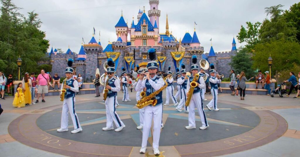 A marching band in white and blue uniforms performs with brass instruments in front of Disneyland’s Sleeping Beauty Castle, surrounded by visitors and colorful decorations.